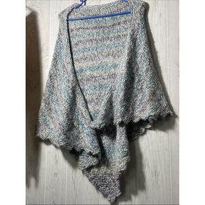 May include: A hand-knitted triangular shawl featuring a variegated yarn pattern in shades of light blue, grey, white, and dark purple. The garment has a textured, chunky knit construction with a scalloped edge along the bottom hem.