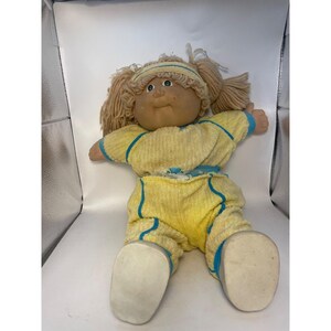 May include: A Cabbage Patch Kids doll with light brown yarn hair styled in pigtails and a matching headband. The doll has a tan vinyl face, green eyes, and wears a yellow textured long-sleeve jumpsuit with blue trim accents and white shoes.