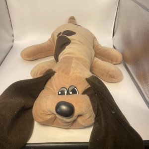 May include: A plush toy dog in a lying down position. The dog is primarily tan with brown patches on its back and ears. It has a black nose and embroidered eyes. The toy is soft and cuddly, designed for children.