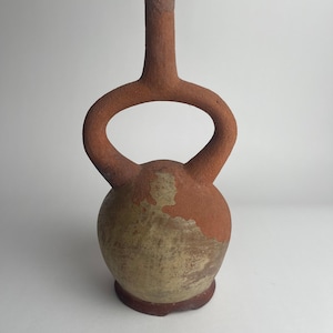 May include: A handcrafted, terracotta-colored ceramic vase with a unique design. The vase features a rounded base, a central bulbous body, and a slender neck with a loop handle. The color is a mix of earthy tones, with a reddish-brown hue and a lighter, mottled surface.
