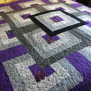 May include: A quilt with a geometric pattern in shades of gray, purple, and black. The quilt is made with a variety of fabrics and has a quilted texture.