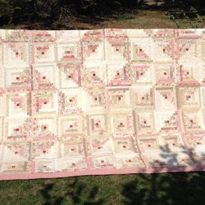 Peut inclure: Une courtepointe rose et crème avec un motif de cabane en rondins. La courtepointe est accrochée à une clôture en bois.