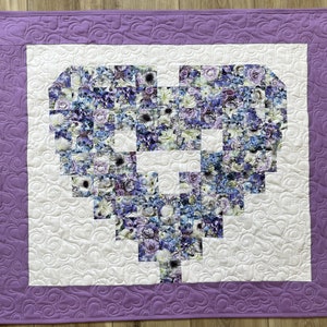 May include: A purple and white quilted wall hanging with a heart-shaped design made of floral fabric squares.