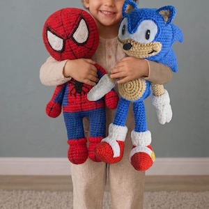 Puede incluir: Dos peluches de ganchillo hechos a mano que representan a un superhéroe rojo y azul con un emblema de araña negra y un erizo azul con guantes blancos, hocico color canela y zapatos rojos, elaborados con puntadas de hilo detalladas.