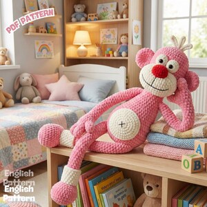 Könnte beinhalten: Ein großer, rosa gehäkelter Affe mit weißem Bauch und roter Nase liegt auf einem Holzregal. Der Affe hat große Augen und lächelt. Auf dem Regal befinden sich auch Bücher und ein Stapel Decken. Die Worte "PDF PATTERN" sind sichtbar.