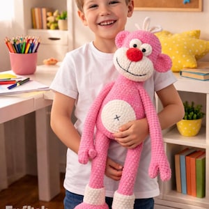 May include: A smiling child holds a large, pink crocheted monkey toy. The monkey has white accents, a red nose, and a white belly with a black cross. The image includes the text "PDF PATTERN" and "English Pattern".