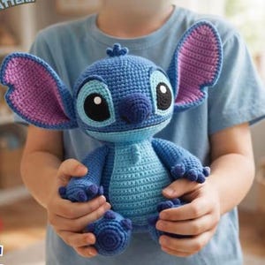May include: A handmade blue crochet Stitch doll with large ears, black eyes, and a light blue belly. The plush toy is held by a person wearing a blue shirt. The doll's ears are lined with purple yarn.