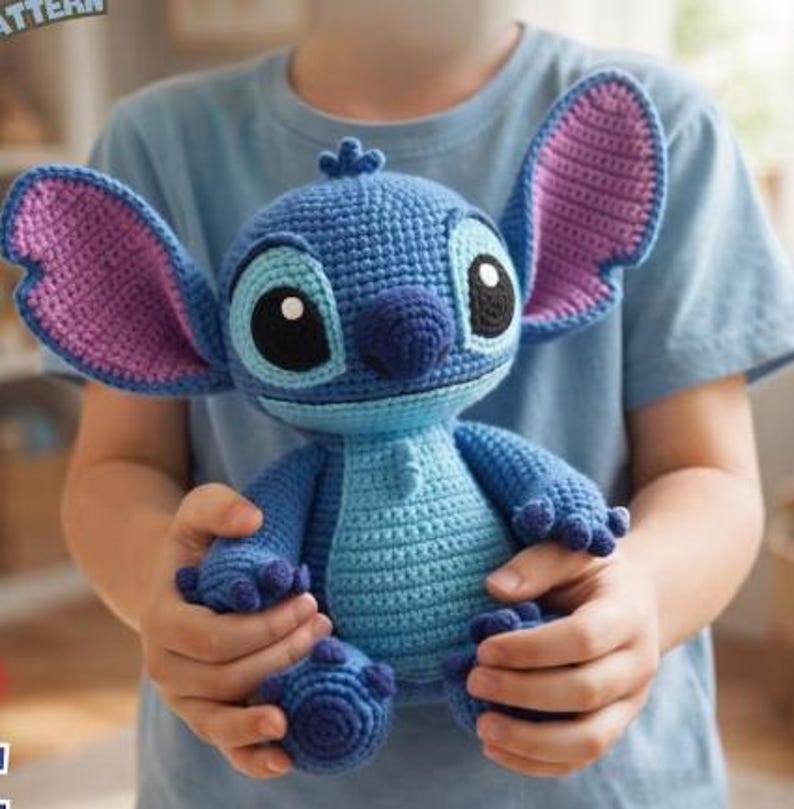 May include: A handmade blue crochet Stitch doll with large ears, black eyes, and a light blue belly. The plush toy is held by a person wearing a blue shirt. The doll's ears are lined with purple yarn.
