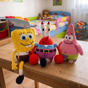 May include: Three crocheted plush toys representing cartoon characters: a yellow rectangular sponge, a red crab with tall eyes, and a pink starfish. The toys feature detailed stitching, vibrant colors, and embroidered facial expressions. Text on the image reads PDF PATTERN and English Pattern.