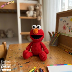 May include: A red crocheted doll featuring a round head, a bright orange nose, large white eyes with black pupils, and a wide black mouth. The doll has a round body with two arms and two legs, all crafted in a consistent red yarn texture. Text reads PDF PATTERN.