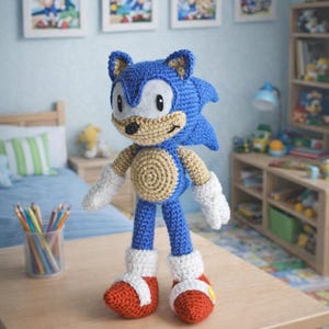 May include: A blue, tan, and white crocheted Sonic the Hedgehog doll stands on a wooden table. The doll has red shoes and white gloves. The image includes framed Sonic artwork and text that reads "PDF Pattern" and "English Pattern".