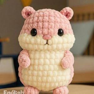 May include: A handmade crochet hamster in shades of pink and cream. The plush toy has black eyes, small ears, and a cute nose. The image includes the text "PDF PATTERN" and "English Pattern".