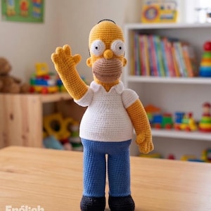 Puede incluir: Una muñeca de ganchillo de un personaje de piel amarilla con cabeza calva, que viste una camisa blanca de cuello y manga corta, pantalones azules y zapatos negros. El personaje tiene una zona bucal bronceada, ojos grandes y un brazo alzado.