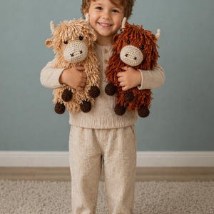 Peut inclure: Un enfant tient deux peluches de vaches des Highlands crochetées. L'une est marron clair, l'autre est marron foncé. Les deux ont des visages crème et de petites cornes. Les peluches ont une fourrure hirsute et sont faites à la main. Les mots "PDF PATTERN" et "English Pattern" sont visibles.