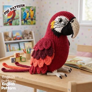 Puede incluir: Un gran loro de ganchillo hecho a mano en tonos rojos, burdeos y naranja, sentado sobre una mesa de madera. El loro tiene un pico blanco y negro y una cabeza color crema. La imagen incluye el texto "PDF PATTERN" y "English Pattern".