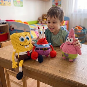 May include: Three crocheted plush toys featuring Spongebob Squarepants, Mr. Krabs, and Patrick Star. The yellow rectangular character, red crab, and pink starfish are handmade with yarn. The image includes the text PDF PATTERN and English Pattern.