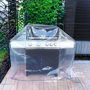 May include: A stainless steel and black outdoor grill covered with a clear plastic cover. The grill has four burners and two black cabinet doors. The cover protects the grill from the elements. The grill is on a wooden deck.