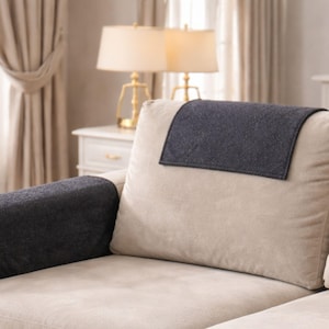 May include: A beige sofa with dark gray armrest covers and a matching headrest cover. The covers appear to be made of a textured fabric. The background includes a white nightstand, gold lamps, and beige curtains.