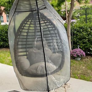 Könnte beinhalten: Ein dunkelbrauner Eierstuhl aus Korbgeflecht mit grauen Kissen ist mit einer transparenten Kunststoffabdeckung mit schwarzem Rand bedeckt. Der Stuhl befindet sich im Freien, mit grünem Laub und einem Baum im Hintergrund.