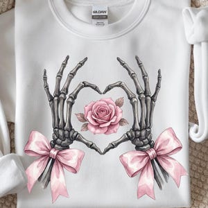 May include: White sweatshirt featuring a graphic of skeleton hands forming a heart around a pink rose. Two pink bows are at the bottom of the design. The sweatshirt has a crew neck and long sleeves.