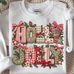 Op de afbeelding: Een wit sweatshirt met ronde hals en de tekst HOLLY JOLLY in een gewatteerd stofpatroon. De letters zijn versierd met rode en groene bloemen-, ruit- en streepmotieven, geaccentueerd met kleine strikjes, belletjes en stervormen.