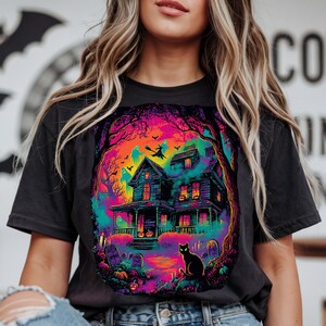 May include: Dark gray t-shirt featuring a vibrant Halloween scene. The graphic depicts a haunted house with a witch flying, ghosts, and a black cat. The design uses bright colors like purple, orange, and teal, creating a spooky yet colorful aesthetic.