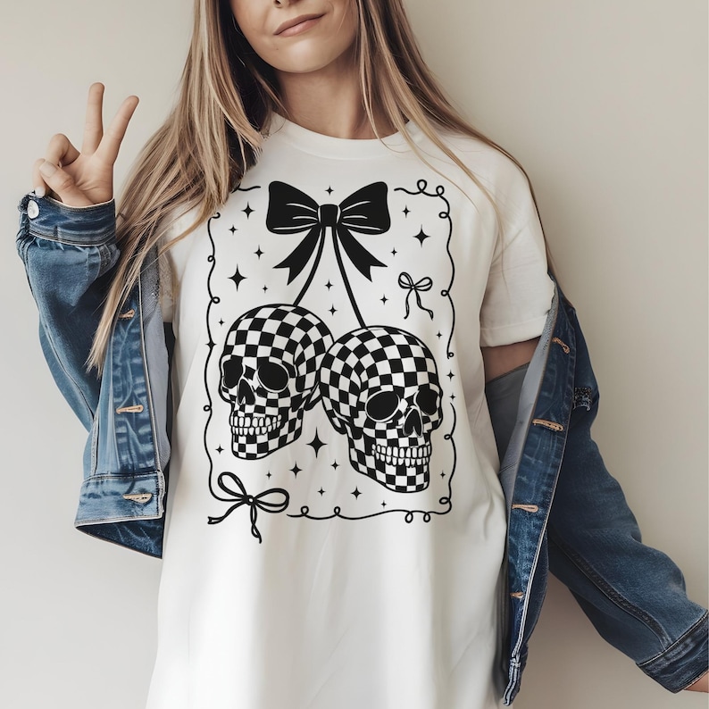 May include: White t-shirt featuring a black and white checkered skull design. The skulls are connected by a bow, with additional bows and stars around the border. The shirt is a casual, everyday wear item.