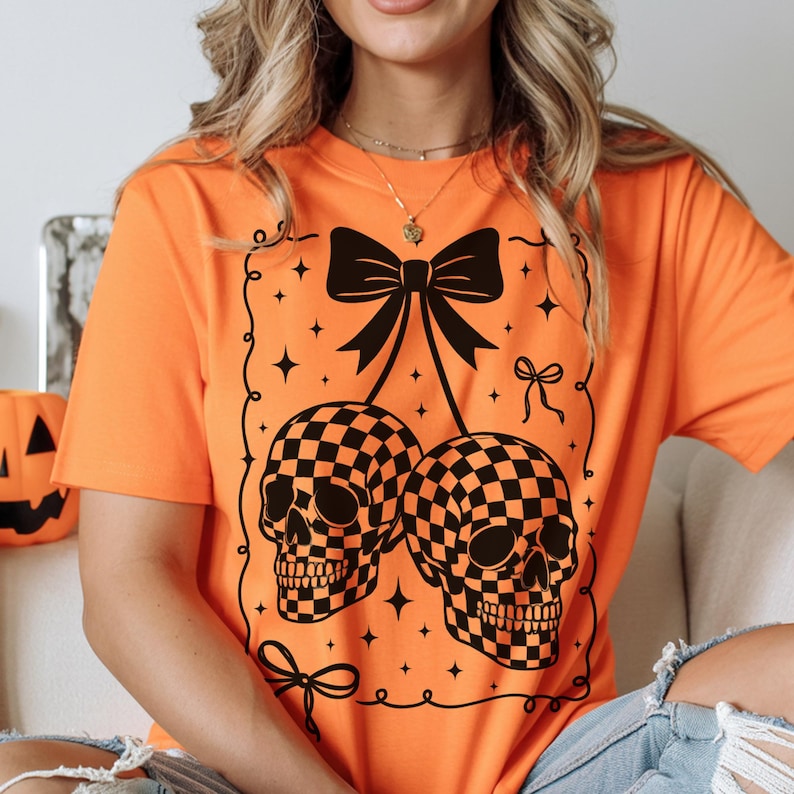 May include: Orange t-shirt featuring a black and white checkered skull design. The graphic includes a black bow, stars, and decorative swirls, creating a Halloween theme. The shirt is short-sleeved.