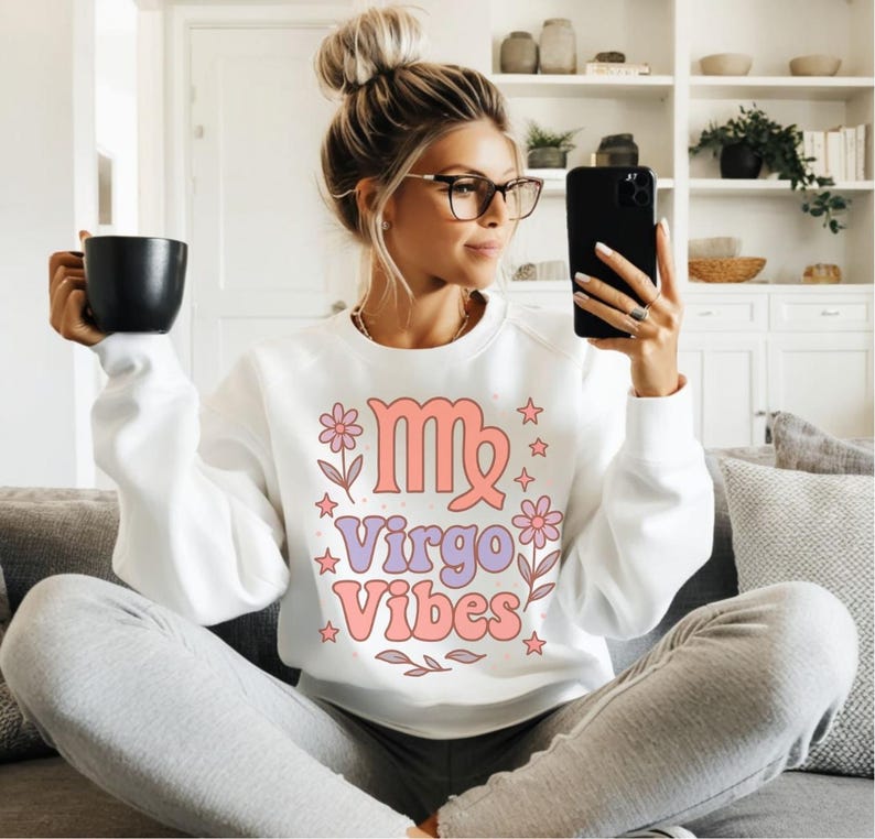 以下が含まれることがあります： 花と星に囲まれたピンクとパープルの乙女座の星座と「Virgo Vibes」の文字が書かれた白いスウェットシャツです。