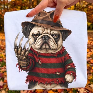 Puede incluir: Una camiseta blanca con una ilustración gráfica de un pug que lleva un sombrero fedora marrón desgastado y un suéter a rayas rojas y verde oscuro hecho jirones. El pug tiene un guante metálico con cuatro cuchillas largas y afiladas en la pata derecha.