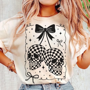 May include: Cream-colored sweatshirt featuring a black and white checkered skull design. The graphic includes two skulls, bows, and stars within a decorative frame. A unique and edgy design for casual wear.