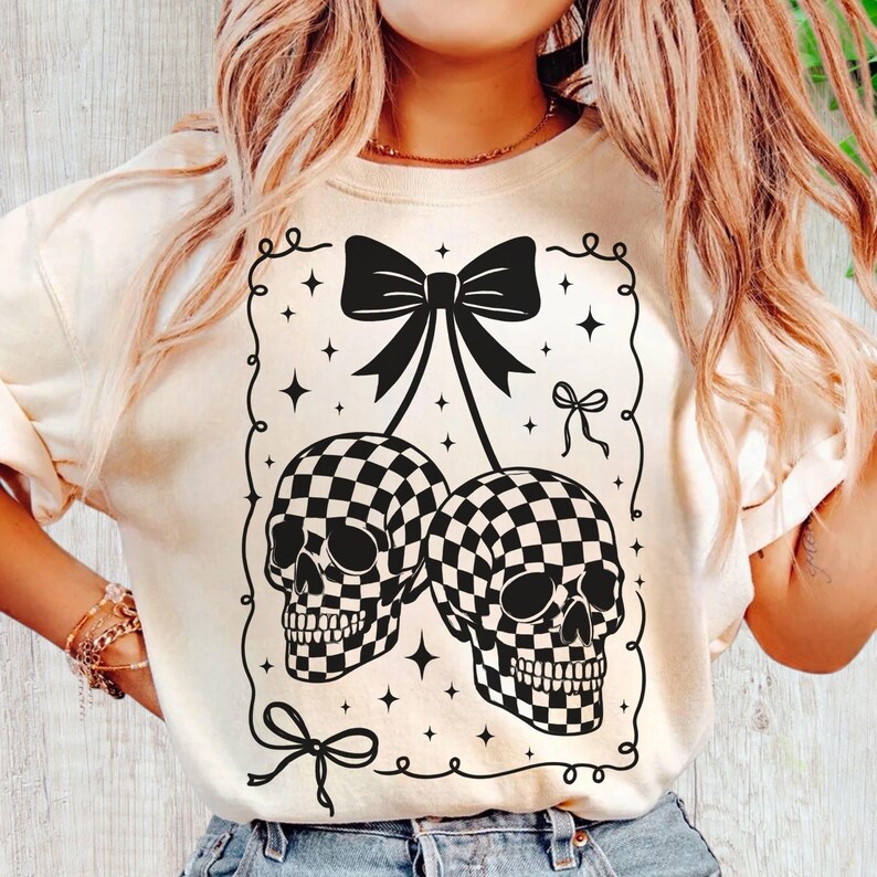 May include: Cream-colored sweatshirt featuring a black and white checkered skull design. The graphic includes two skulls, bows, and stars within a decorative frame. A unique and edgy design for casual wear.