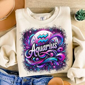 May include: A cream-coloured sweatshirt with a colourful graphic design featuring the zodiac sign Aquarius. The design includes a blue and pink wave symbol, the word "Aquarius" in a stylised font, and a background of purple, pink, and blue swirls.