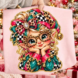 May include: A light pink sweatshirt featuring a graphic illustration of an elf wearing leopard print glasses, a matching hat with a large bow, and a collar adorned with gold bells. The elf has large green eyes and is resting their chin on their hands.