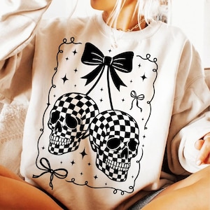 May include: Cream-coloured sweatshirt with a black and white chequered skull design. The graphic includes two skulls, a large bow, and decorative stars. The design is outlined with a black border.
