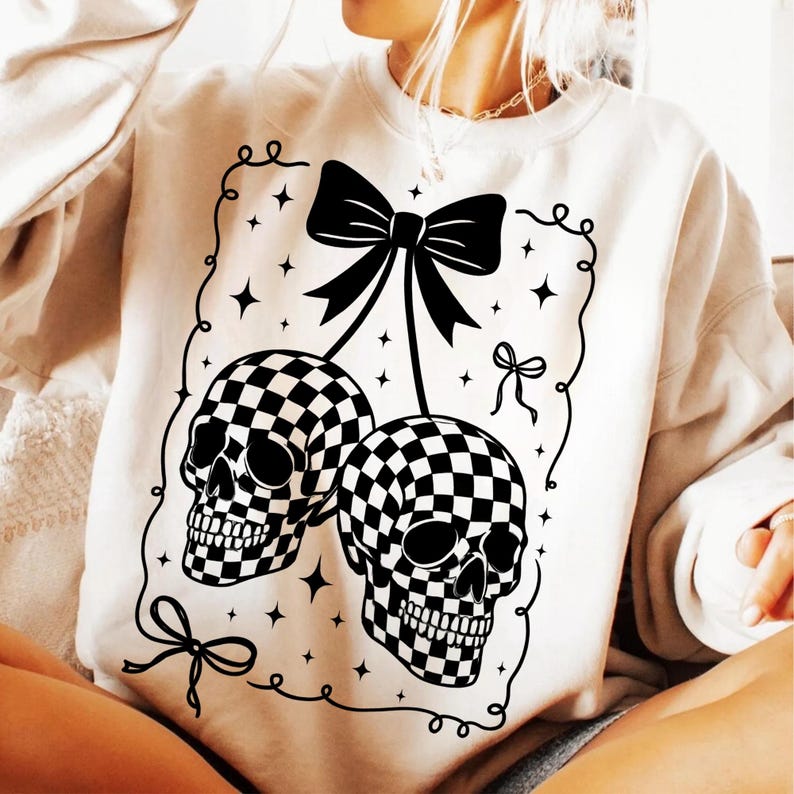 May include: Cream-colored sweatshirt featuring a black and white checkered skull design. The graphic includes two skulls, a large bow, and decorative stars. The design is outlined with a black border.