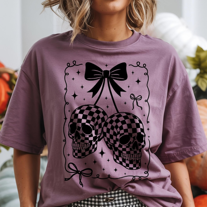 May include: A mauve-colored t-shirt featuring a black graphic of two skulls designed as cherries with a checkered pattern. The skulls are connected by a black bow, with additional bows and stars in the design. The shirt has short sleeves.