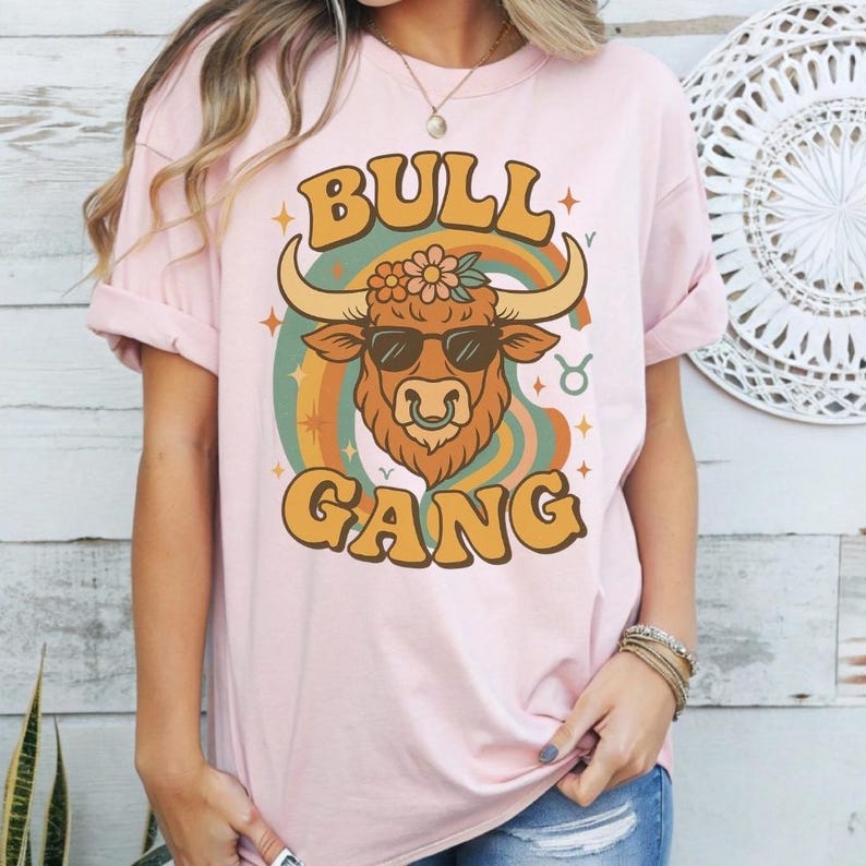 以下が含まれることがあります： ピンクのTシャツに、サングラスと鼻ピアスをした牡牛のレトロなグラフィックがプリントされています。グラフィックには、カラフルなレトロなフォントで「Bull Gang」と書いてあります。
