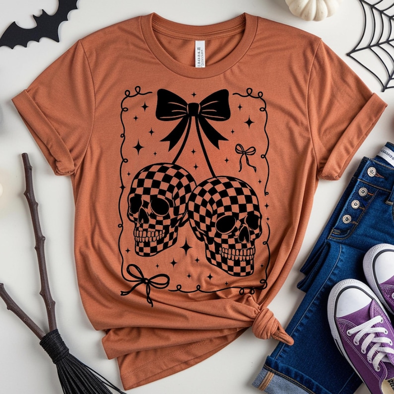 May include: Rust-colored t-shirt featuring a black graphic of two checkered skull heads with a bow and decorative flourishes. The shirt is styled with rolled sleeves and a knot at the waist, paired with blue jeans and purple sneakers.