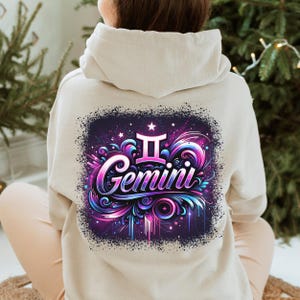 May include: A light beige hoodie featuring a graphic print of the Gemini zodiac sign. The graphic is in a colourful style with pink, blue, and purple hues. The sign is surrounded by swirls and stars.