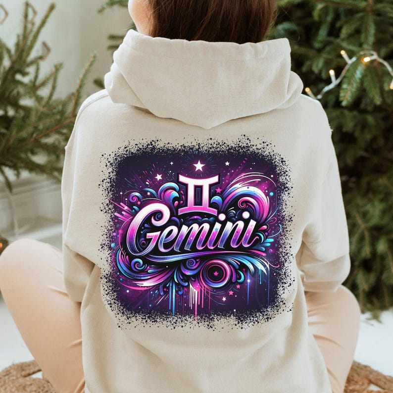 May include: A light beige hoodie featuring a graphic print of the Gemini zodiac sign. The graphic is in a colourful style with pink, blue, and purple hues. The sign is surrounded by swirls and stars.