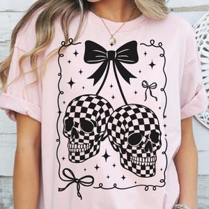 May include: A light pink t-shirt featuring a black and white checkered skull design. The skulls are connected by a black bow, with additional bow and star details. The shirt has short sleeves and a relaxed fit.