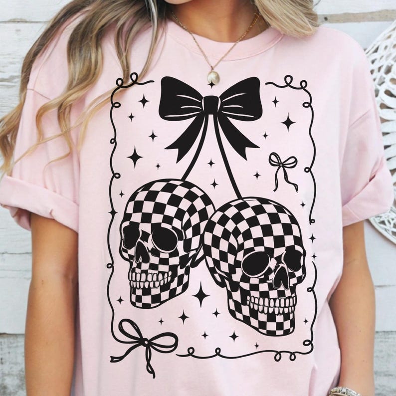 May include: A light pink t-shirt featuring a black and white checkered skull design. The skulls are connected by a black bow, with additional bow and star details. The shirt has short sleeves and a relaxed fit.