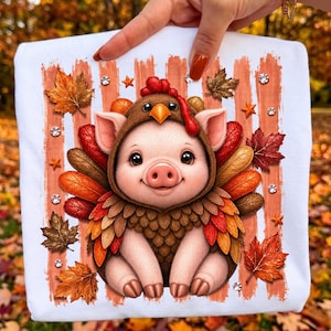 Peut inclure: Un t-shirt blanc avec un motif représentant un porcelet rose portant un costume de dinde avec des plumes brunes et orange. Le design inclut des feuilles d'automne, des petites étoiles orange et des strass scintillants sur des rayures verticales.