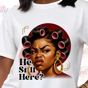 Pode incluir: Camiseta branca com um gráfico de uma mulher com rolos e brincos de argola dourados, com o texto "He Still Here?". O retrato da mulher está sobre um círculo vermelho. A camiseta tem mangas curtas.