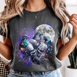 May include: Dark gray t-shirt featuring a graphic of three pigeons with iridescent feathers, set against a backdrop of a full moon and a starry night sky. The design incorporates shades of purple, blue, and white.