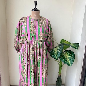 Könnte beinhalten: Ein langes, fließendes Kleid mit einem einzigartigen Streifenmuster. Das Kleid hat abwechselnd vertikale Streifen in leuchtendem Pink und Grün mit einem Tigerdruck-Design. Das Kleid hat lange Ärmel und eine Krawatte am Ausschnitt, präsentiert auf einer Schaufensterpuppe.