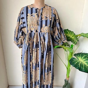 Könnte beinhalten: Ein langes Kleid mit Tigerstreifenmuster in Schwarz und Gold auf hellblauem Hintergrund. Das Kleid hat einen runden Ausschnitt mit Bindeband und lange, puffärmelige Ärmel. Das Kleid wird auf einer Schaufensterpuppe präsentiert.