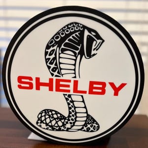 May include: A white circular sticker with a black outline. The sticker features a black and white cobra with the word "SHELBY" in red letters.