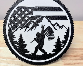 May include: Circular black and white metal art piece depicting Bigfoot holding a flag and a stick, with a mountain range and American flag design. The outer edge has a decorative, ridged border.